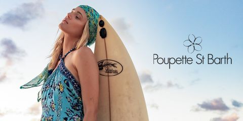 Eine Person in einem blauen Blumenoutfit und Kopftuch steht vor einem Surfbrett. Der Himmel ist mit weichen Wolken bedeckt. Rechts ist der Text „Poupette St Barth“ mit einem Blumenlogo zu sehen.