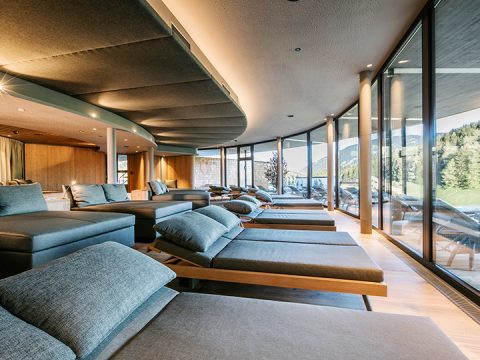 Eine moderne Entspannungslounge mit großen Fenstern, die einen malerischen Blick auf Bäume und Berge bieten. Der Raum verfügt über mehrere gepolsterte Loungesessel mit sanfter Beleuchtung, die eine ruhige Atmosphäre schafft.