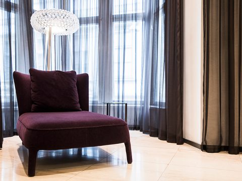 Ein moderner Raum mit einem weichen, dunkelvioletten Sessel und einem passenden Kissen. Daneben stehen ein eleganter Beistelltisch und eine moderne Lampe mit strukturiertem Schirm. Vom Boden bis zur Decke reichende Fenster mit durchsichtigen schwarzen Vorhängen lassen natürliches Licht herein. Marmorboden rundet den Look ab.