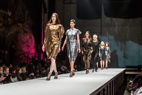 Eine Gruppe von Models läuft in metallischen Kleidern in Gold- und Silbertönen über einen Laufsteg. Das Publikum sitzt auf beiden Seiten und im Hintergrund ist eine schwach beleuchtete Industriekulisse zu sehen.