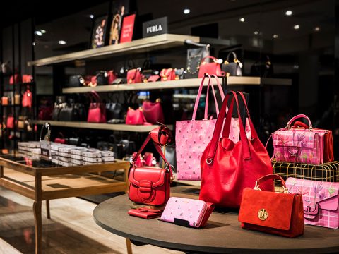 Eine Ausstellung von Luxushandtaschen in einem Geschäft, mit verschiedenen Stilen in Rot- und Rosatönen. Im Hintergrund sind Regale mit weiteren Handtaschen und Accessoires bei sanfter Beleuchtung zu sehen.