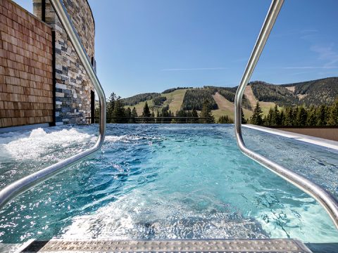 Blick von einem Außenpool mit Metallhandläufen, die in das klare, blaue Wasser führen. Um den Pool herum sind malerische Berge und Pinien unter einem klaren, blauen Himmel zu sehen.