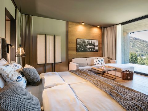 Ein gemütliches Hotelzimmer mit großem Bett, moderner Holzvertäfelung und sanften Farbtönen. Ein Sofa und ein Couchtisch stehen neben einem Fenster mit Blick auf die Berge. Elegante Beleuchtung und Dekoration sorgen für eine warme Atmosphäre.