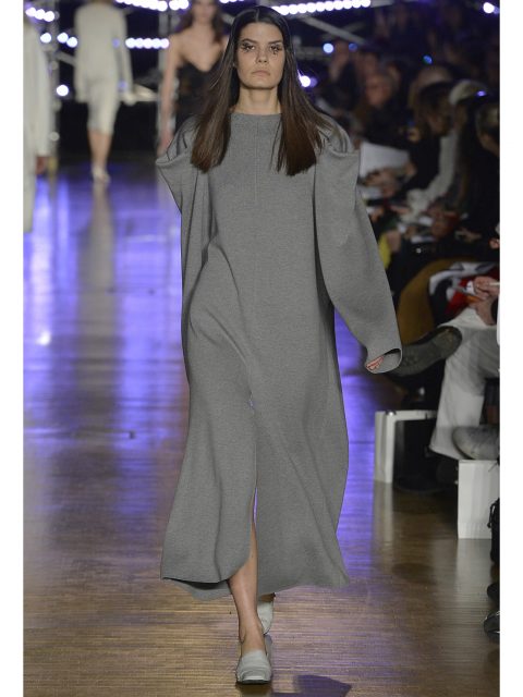 Ein Model läuft in einem langen, grauen Kleid mit weiten Ärmeln über den Laufsteg. Im Hintergrund sind ein sitzendes Publikum und helle Laufsteglichter zu sehen.