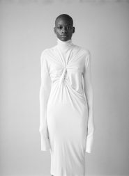 Eine Person steht vor einem neutralen Hintergrund und trägt ein langärmeliges, weißes Kleid mit hohem Kragen und Knotendetail auf der Brust. Das Kleid reicht bis zur Mitte der Wade. Der Gesichtsausdruck der Person ist ruhig und direkt.