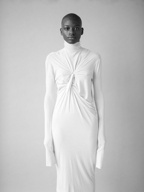 Eine Person steht vor einem neutralen Hintergrund und trägt ein langärmeliges, weißes Kleid mit hohem Kragen und Knotendetail auf der Brust. Das Kleid reicht bis zur Mitte der Wade. Der Gesichtsausdruck der Person ist ruhig und direkt.