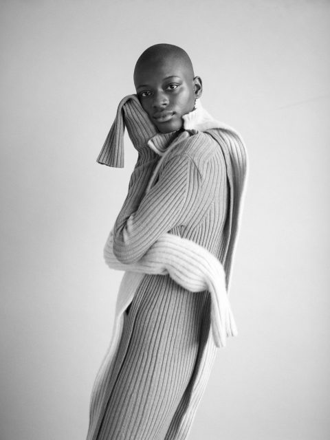 Eine Person posiert selbstbewusst in einem gemütlichen, langen Rippenstrickpullover mit passendem Schal. Das Bild ist schwarzweiß und hebt die Textur des Outfits hervor. Der Hintergrund ist einfach und neutral und lenkt den Fokus auf die Person.