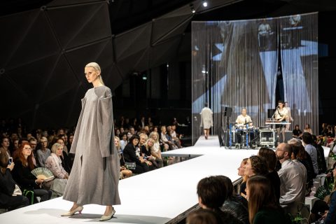 Ein Model in einem langen, grauen Mantel läuft über einen Laufsteg, umgeben von einem Publikum. Im Hintergrund sind Musiker zu sehen. Der Laufsteg und die Kulisse haben ein modernes, geometrisches Design.