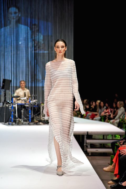 Ein Model läuft in einem langen, halbtransparenten, gestreiften Kleid über einen hell erleuchteten Laufsteg. Im Hintergrund sind ein Musiker am Schlagzeug und ein Publikum entlang der Bühne zu sehen.