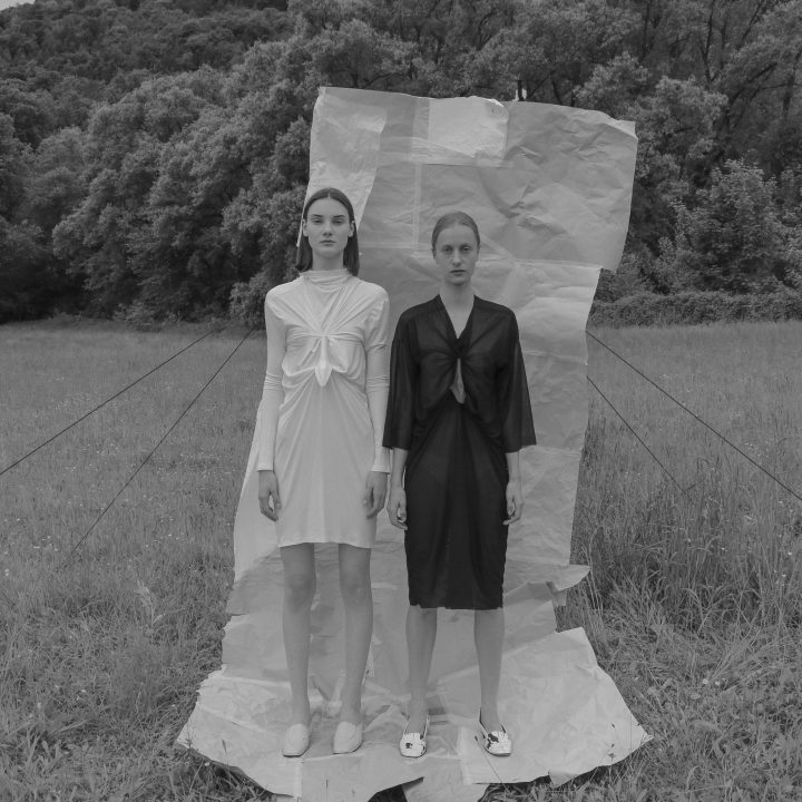 Zwei Frauen stehen auf einer Wiese vor einem Hintergrund aus zerknülltem Papier. Eine trägt ein helles Kleid, die andere ein dunkles. Beide Kleider haben ähnliche Knotenmuster. Im Hintergrund sind Bäume und Hügel zu sehen. Das Bild ist schwarzweiß.