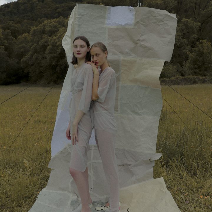 Zwei Personen stehen vor einem großen, zerknüllten Papierhintergrund auf einem Feld im Freien. Beide tragen halbtransparente, helle Kleidung. Im Hintergrund sind Bäume und Hügel unter einem bewölkten Himmel zu sehen.
