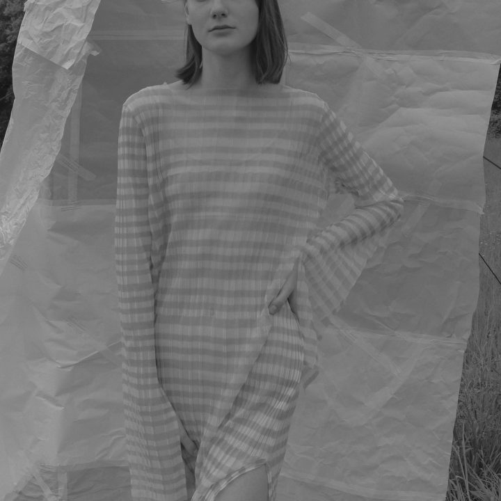 Ein Schwarzweißfoto einer Person in einem langen, karierten Kleid, die im Freien steht. Sie hat eine Hand auf der Hüfte, hinter ihr liegt ein großes Blatt zerknülltes Papier, das einen strukturierten Hintergrund bildet.