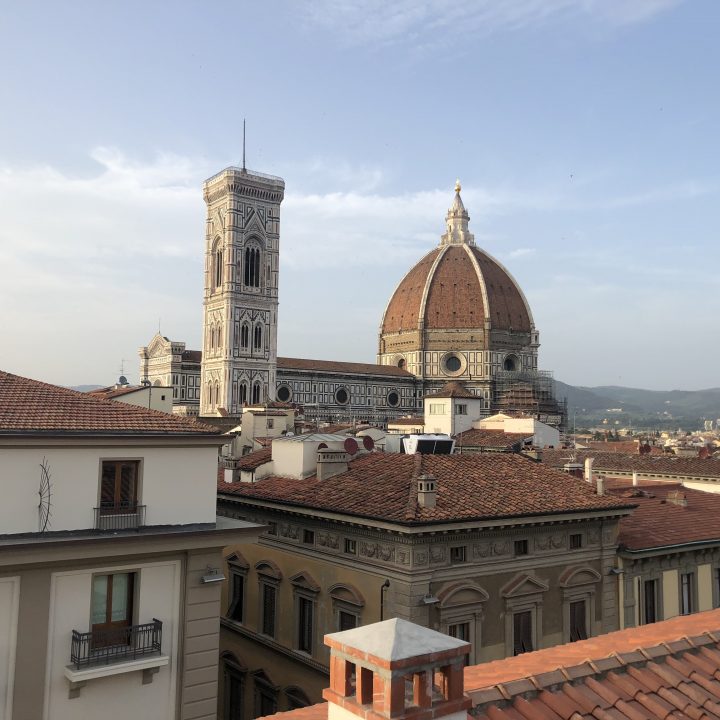 Blick vom Dach von Florenz, Italien, auf die Kathedrale von Florenz mit ihrer ikonischen Kuppel und dem Glockenturm. Im Vordergrund sind traditionelle Terrakotta-Dächer unter einem klaren blauen Himmel zu sehen.