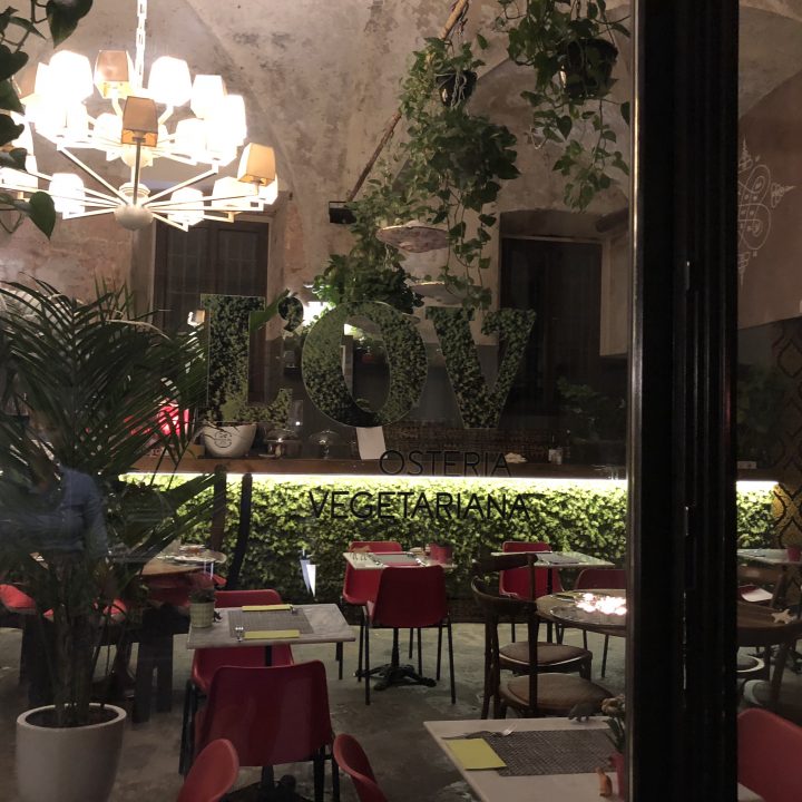 Ein gemütliches, schwach beleuchtetes vegetarisches Restaurant mit roten und weißen Stühlen, grünen Pflanzen und einem großen Kronleuchter. Im Fenster steht der Text „LOVE OSTERIA VEGETARIANA“.