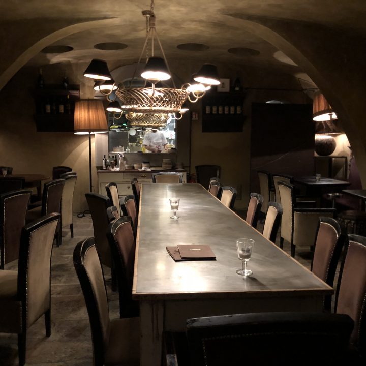 Ein schwach beleuchtetes, gemütliches Restaurant-Interieur mit einem langen Tisch, umgeben von Stühlen. Warmes Licht von Kronleuchtern und Stehlampen schafft eine intime Atmosphäre. Auf dem Tisch liegen ein Buch und ein paar Gläser, im Hintergrund ist rustikales Dekor zu sehen.