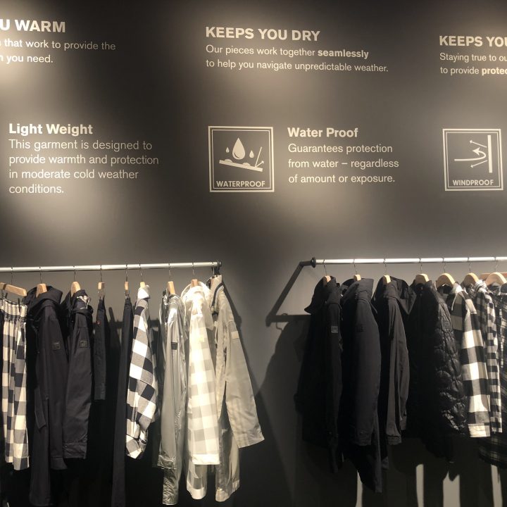 Kleiderständer mit verschiedenen Mänteln und Jacken. An der Wand sind Grafiken und Texte angebracht, die die Eigenschaften Wärme, geringes Gewicht, Wasser- und Windfestigkeit hervorheben.