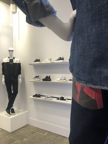 Innenansicht eines Einzelhandelsgeschäfts mit einer Schaufensterpuppe in schwarz-weißem Outfit (links). In den Regalen sind verschiedene Turnschuhe und Accessoires ausgestellt. Im Vordergrund trägt eine weitere Schaufensterpuppe eine Jeansjacke.
