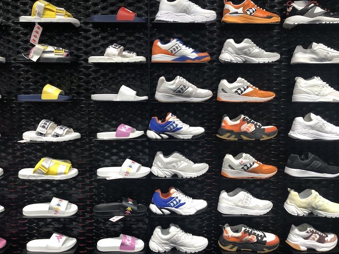Eine Ausstellung verschiedener Turnschuhe und Pantoletten an einer schwarzen Wand. Die Kollektion umfasst verschiedene Stile und Farben wie Weiß, Orange, Gelb und Blau. Jedes Regal enthält verschiedene Designs und zeigt die verfügbare Vielfalt.