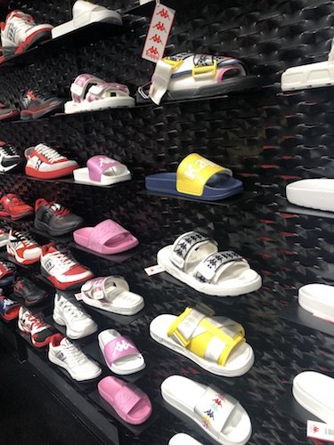Eine Schauwand mit verschiedenen Sportschuh- und Sandalenarten in unterschiedlichen Farben, darunter Rot, Rosa, Gelb und Blau. Die Schuhe sind ordentlich auf dunklen Regalen in einem Geschäft angeordnet.