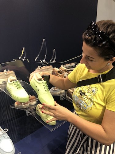 Eine Frau in einem gelben Shirt mit Tiger-Print und einer Sonnenbrille auf dem Kopf betrachtet ein Paar hellgrüne Sneakers auf einem Ausstellungsregal mit mehreren anderen Schuhen.