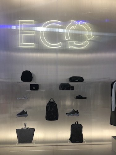 Eine moderne Präsentation umweltfreundlicher Modeartikel, darunter Taschen, Schuhe und Accessoires, ordentlich auf transparenten Regalen angeordnet. Darüber wird das Wort „ECO“ mit Recyclingpfeilen beleuchtet, was das Nachhaltigkeitsthema hervorhebt.