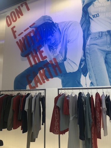 Ein Schaufenster in einem Bekleidungsgeschäft mit einem großen Bild von zwei Models in legerer Kleidung. An der Wand steht in fetten roten Buchstaben der Text „DON’T F--- WITH THE EARTH“. Darunter sind Kleiderständer mit Kleidung in verschiedenen Farben ordentlich angeordnet.