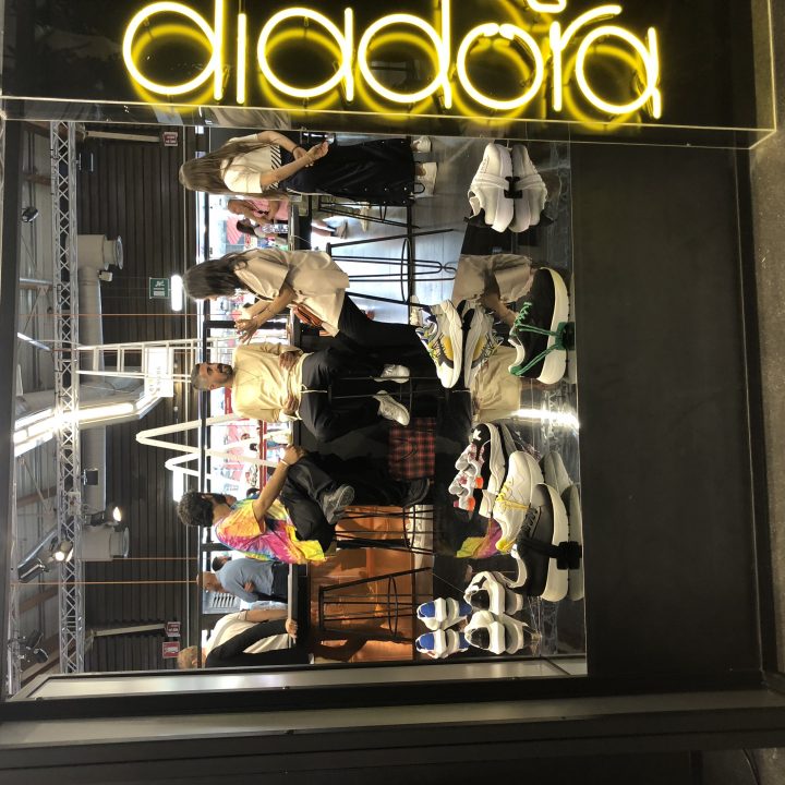 Menschen stöbern und probieren Turnschuhe in einem Diadora-Geschäft aus. Verschiedene bunte Schuhe sind auf Regalen und einem Tisch angeordnet. Oben im Fenster ist ein neongelbes „Diadora“-Schild zu sehen. Das Geschäft hat ein modernes, industrielles Interieur.
