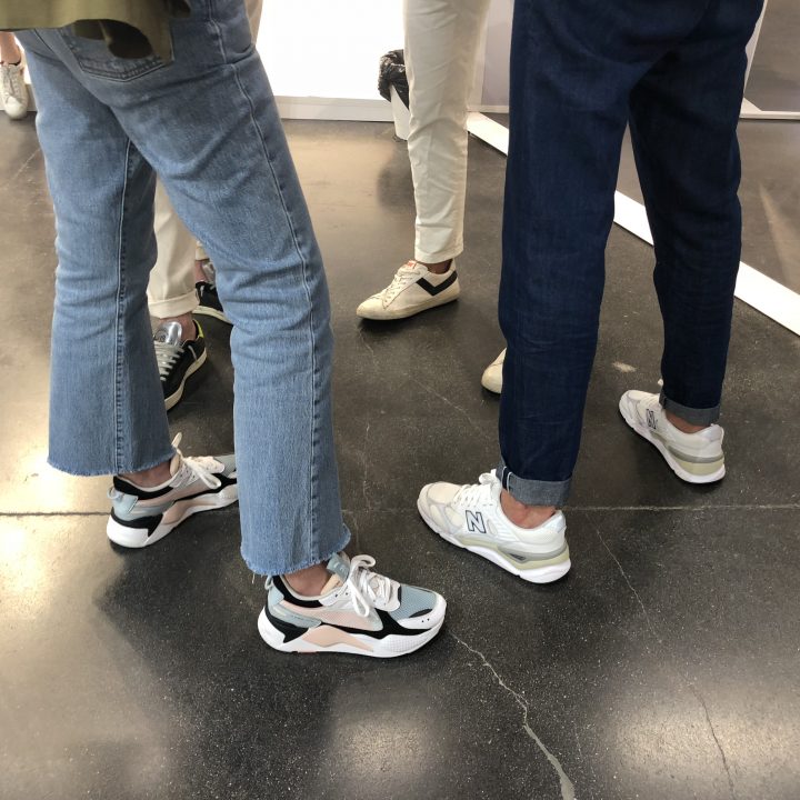 Eine Gruppe von Menschen steht in einem Raum und trägt lässige Turnschuhe und Jeans. Der Fokus liegt auf ihren Schuhen und Unterschenkeln vor einem dunklen Boden.
