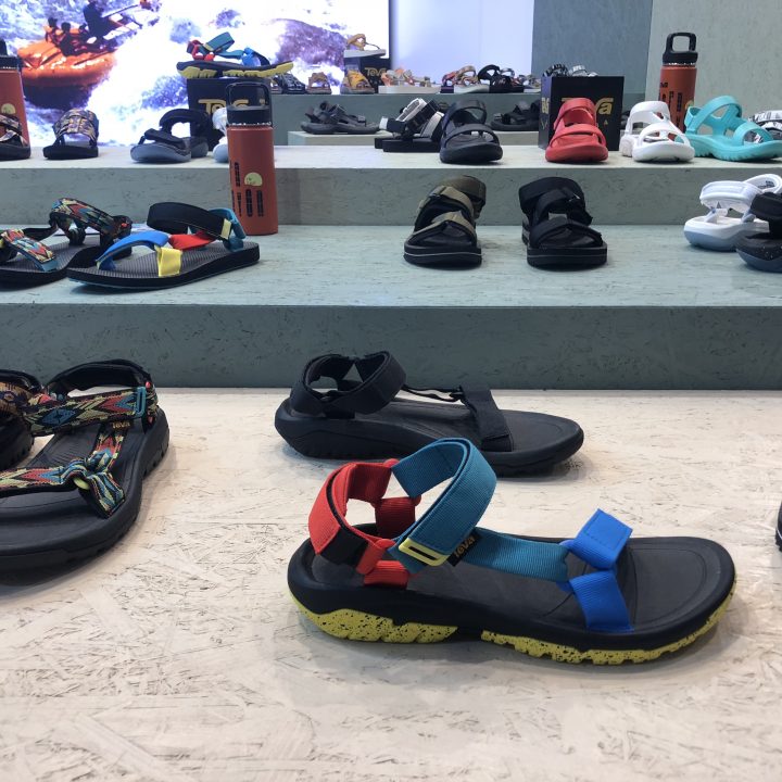 Eine Ausstellung bunter Sandalen auf Stufen. Verschiedene Stile und Designs, darunter Sandalen mit bunten Riemen und dicken Sohlen, werden präsentiert. Zwischen den Sandalen stehen ein paar Wasserflaschen.
