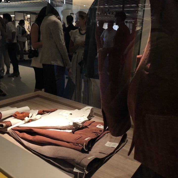 Eine Vitrine mit ordentlich gefalteten Kleidungsstücken in Braun- und Weißtönen. Im Hintergrund laufen und interagieren Menschen, was auf eine belebte Ausstellungs- oder Ladenumgebung hindeutet.