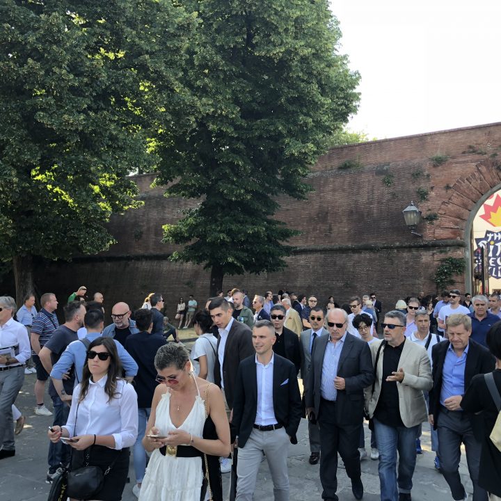 Eine große Gruppe von Menschen geht draußen in der Nähe einer Backsteinmauer und eines gewölbten Eingangs spazieren. In der Nähe stehen Bäume. Einige tragen formelle Kleidung wie Anzüge und Kleider. Über dem Eingang ist eine Flagge zu sehen.