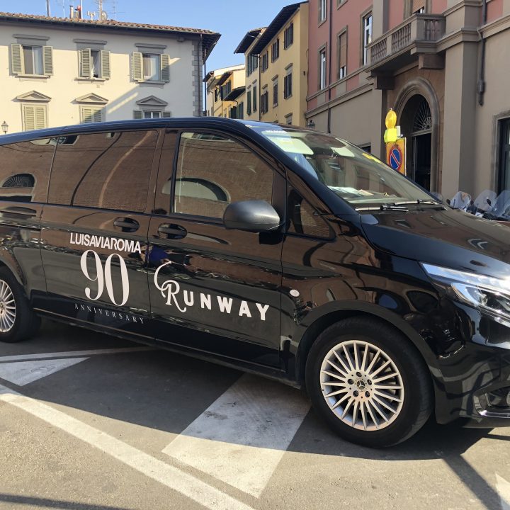 Ein schwarzer Mercedes-Benz-Transporter steht auf einer Straße. Auf seiner Seite prangt der Schriftzug „LuisaViaRoma 90th Anniversary Runway“. Er ist von historischen Gebäuden umgeben und im Hintergrund sind an einem sonnigen Tag ein paar Menschen zu sehen.