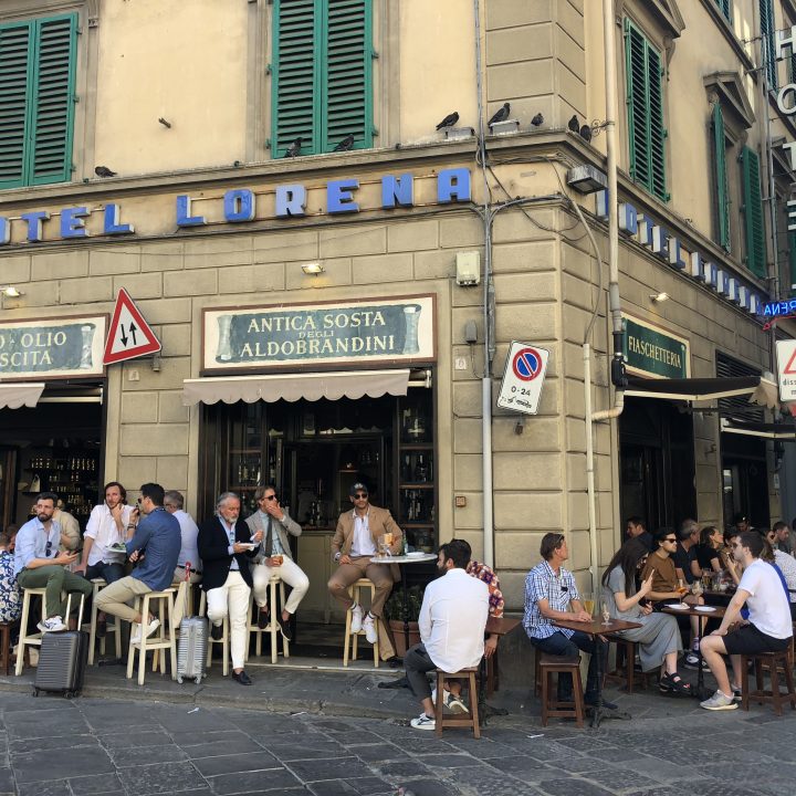 Eine geschäftige Straßencafé-Szene an einer Straßenecke vor dem Hotel Lorena. Leute sitzen an Tischen unter Schildern mit der Aufschrift „Vino - Olio Mescita“ und „Antica Sosta Aldobrandini“. Die Straße ist voller Fußgänger und parkender Motorroller.