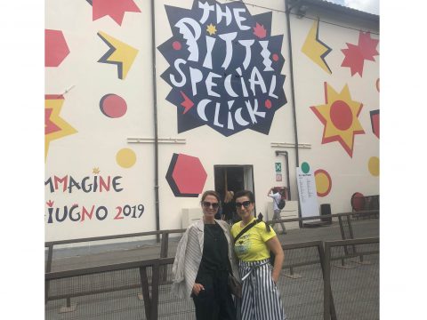 Zwei Personen stehen vor einer bunten Wand mit geometrischen Formen und den Worten „The Pitti Special Click“. Auf der Wand sind außerdem italienische Texte und ein Datum aus dem Jahr 2019 zu sehen. Beide Personen lächeln und tragen legere Kleidung.