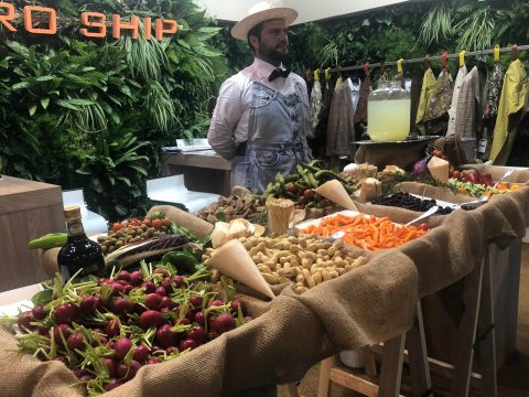 Ein Mann mit Strohhut und Overall steht hinter einem rustikalen Marktstand voller frischer Produkte, darunter Radieschen, Nüsse, Paprika und Karotten. Auf dem Tisch steht ein Krug Limonade. Im Hintergrund sind üppiges grünes Laub und Kleidung ausgestellt.