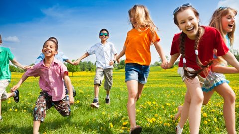 Kinder halten sich an den Händen und laufen fröhlich durch ein Feld mit gelben Blumen unter einem klaren blauen Himmel. Sie sind in farbenfrohe Sommerkleidung gekleidet und einige tragen Sonnenbrillen und genießen den sonnigen Tag.