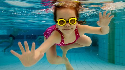 Ein kleines Kind schwimmt unter Wasser in einem Pool. Es trägt eine gelbe Schwimmbrille und einen rosa gepunkteten Badeanzug. Die Hände des Kindes sind ausgestreckt und um sie herum steigen Blasen auf. Im Hintergrund sind andere Schwimmer zu sehen.