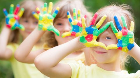 Drei Kinder in gelben Hemden stehen in einer Reihe im Freien. Jedes hält spielerisch seine bunt bemalten Hände vors Gesicht und bildet mit seinen Fingern einen Rahmen. Der Hintergrund ist eine verschwommene grüne Umgebung, die eine spielerische Aktivität im Freien suggeriert.