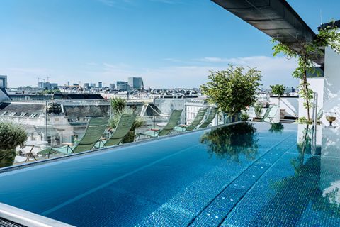 Infinity-Pool auf dem Dach mit klarem blauen Himmel und Stadtlandschaft im Hintergrund. Mehrere grüne Liegestühle sind auf dem Deck angeordnet und das Grün verleiht einen Hauch von Natur. Die Szene bietet einen Panoramablick auf städtische Gebäude.