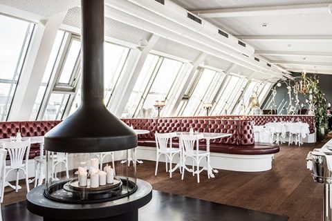 Ein modernes Restaurant-Interieur mit einem runden Kamin mit Kerzen im Vordergrund. Der Raum umfasst rot gepolsterte Sitzbänke an großen Fenstern, weiße Stühle und Tische mit Holzboden und elegantem Dekor.