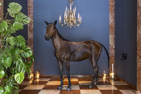 Eine realistische Skulptur eines Pferdes, das in einem eleganten Raum mit Marmorsäulen und einer dunkelblauen Wand steht. Darüber hängt ein Kronleuchter, und im Raum sind Kerzen verteilt. Auf der linken Seite sind üppige grüne Pflanzen zu sehen.