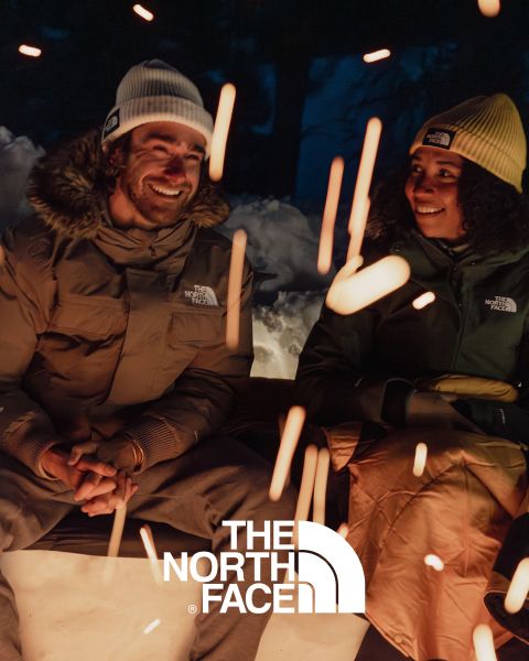 Zwei Menschen sitzen am Lagerfeuer und lächeln sich an. Sie tragen Jacken und Beanies von North Face. Im Vordergrund sind Funken vom Feuer zu sehen. Die Szene spielt vor einer verschneiten Nachtkulisse mit dem Logo von North Face am unteren Bildrand.