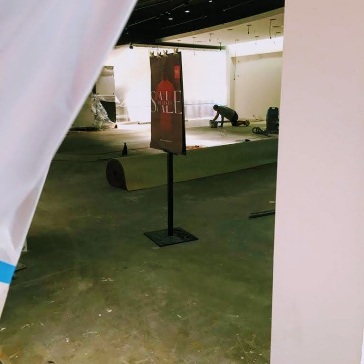 Ein teilweise fertiggestellter Verkaufsraum mit einem „SALE“-Schild auf einem Ständer. Im Hintergrund ist ein Arbeiter auf dem Boden zu sehen, der wahrscheinlich Material ausrollt. Der Bereich ist schwach beleuchtet und es gibt unfertige Wände und Böden.
