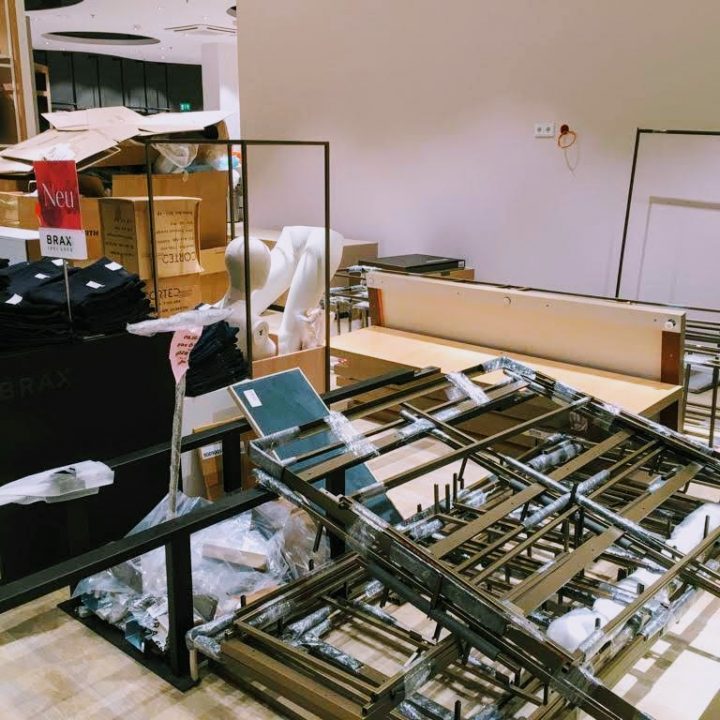 Ein unordentlicher Ladenbereich mit Kartons, gefalteten Kleidungsstücken und Metallgestellen, die auf dem Boden verstreut sind. Links ist ein rotes Schild mit der Aufschrift „Neu“ zu sehen, umgeben von unverpackten Materialien und teilweise montierten Einrichtungsgegenständen.