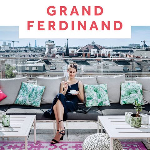 Eine Frau in einem schwarzen Kleid sitzt auf einem Sofa auf einer Dachterrasse mit Kissen im Tropen-Stil und hält eine Tasse in der Hand. Im Hintergrund erstreckt sich die Skyline der Stadt. Darüber ist der Text „Grand Ferdinand“ zu sehen.