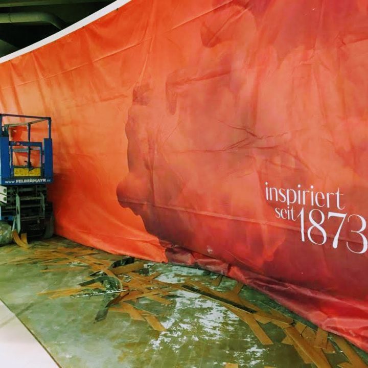 Eine blaue Scherenhebebühne steht neben einer großen, teilweise angebrachten roten Wandverkleidung mit dem deutschen Text „inspiriert seit 1873“. In der Nähe liegen Holzbretter und Schutt verstreut auf dem Boden.