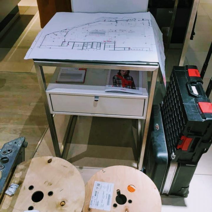 Auf einem kleinen Tisch mit einer Schublade liegt ein Bauplan. In der Nähe stehen zwei Holzspulen mit Elektrokabel und ein großer schwarzer Werkzeugkasten. Die Szene scheint ein Arbeits- oder Baubereich im Innenbereich zu sein.