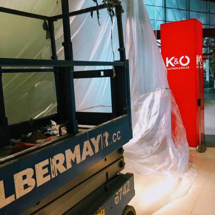 Eine Baustelle in einem Gebäude mit einem blauen Aufzug, auf dem das Wort „ALBERMAYER“ zu sehen ist. Eine Plastikplane hängt herunter und bedeckt den Bereich teilweise. In der Nähe steht auf einer roten Säule „K&O“ in weißen Buchstaben.