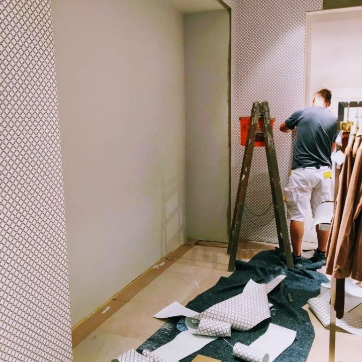 Ein Mann steht auf einer Leiter und tapeziert eine Wand. Die Fläche ist mit Schutzfolien und abgerollter Tapete bedeckt. Die Wände sind teilweise mit einem geometrischen Muster bedeckt und auf dem Boden liegen verschiedene Werkzeuge verstreut.