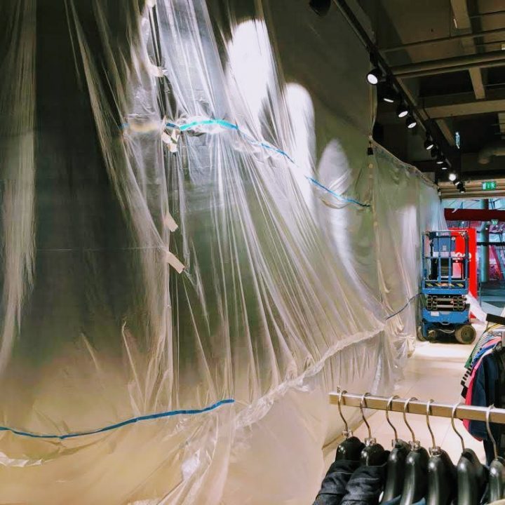 Ein Einzelhandelsgeschäft im Umbau, dessen große Bereiche mit transparenten Plastikfolien verkleidet sind. Im Vordergrund sind Kleider auf Kleiderbügeln zu sehen. Deckenlampen beleuchten die Szene, im Hintergrund ist ein Palettenwagen zu sehen.